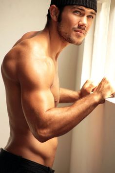 a shirtless man with no shirt on leaning against a window sill looking at the camera