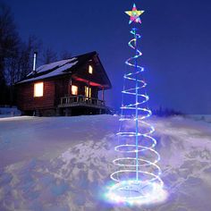 a lit up christmas tree in the snow near a cabin at night with lights on it