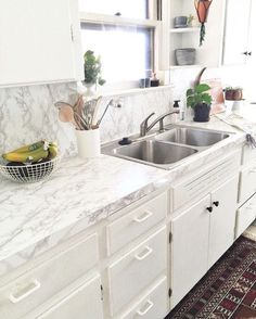 a kitchen with white cabinets and marble counter tops, plants in the window sill