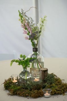 an arrangement of flowers and greenery is displayed in a glass vase on a moss covered table