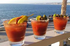 three glasses filled with different types of drinks on a table near the ocean and beach