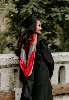 a woman in graduation gown leaning against a wall and looking off into the distance with trees behind her