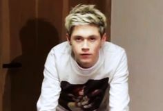 a young man with blonde hair sitting on a stool in front of a door and looking at the camera