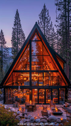 An illuminated A-frame cabin Airbnb nestled in the mountains at twilight, surrounded by towering pine trees. The warm glow from the interior lights creates a cozy and inviting atmosphere, with an outdoor fire pit and seating area set against a stunning forest backdrop.