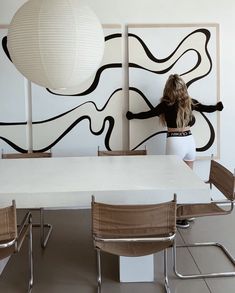 a woman standing in front of a white table with four chairs and a large painting on the wall behind her