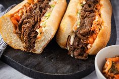 two sandwiches with meat, cheese and sauce on a cutting board next to a bowl