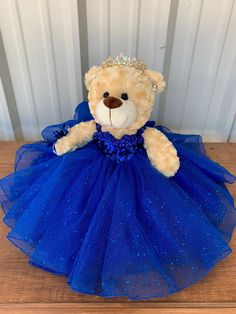 a teddy bear wearing a blue dress on a wooden table next to a white wall