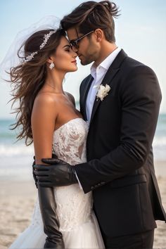 a bride and groom standing on the beach with their arms around each other, wearing black gloves