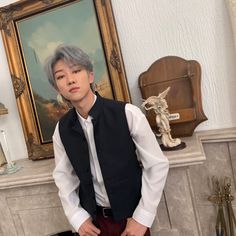 a man in a black vest and white shirt standing next to a fireplace with a painting on the wall behind him