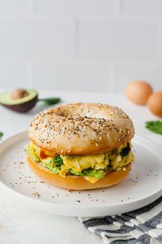 a bagel sandwich on a plate with an avocado in the background