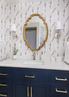 a bathroom vanity with blue cabinets and gold trim on the mirror, along with wallpaper