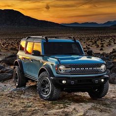 a blue truck parked on top of a dirt field next to rocks and mountains at sunset