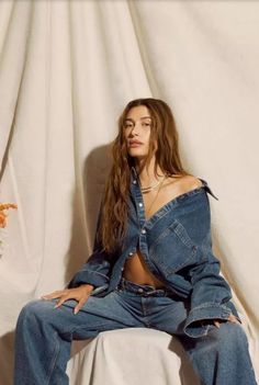 a woman sitting on top of a white bed next to a curtain with her hands in her pockets