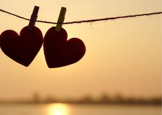 two red hearts hanging from a line with the sun setting in the background and water behind them