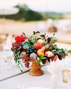 an arrangement of fruit and flowers on a table with wine glasses, plates and napkins