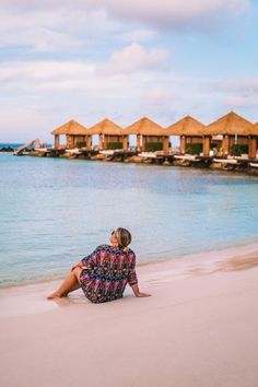 a woman sitting on the beach in front of some thatched huts and looking at the water