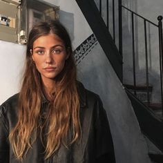 a woman with long hair standing in front of a stair case and looking at the camera