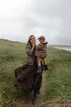 a woman carrying a child on her back while walking through tall grass near the ocean