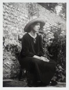 an old black and white photo of a woman wearing a hat sitting in front of a stone wall