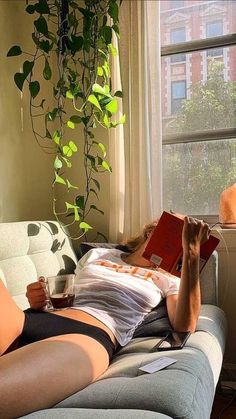 a woman laying on a couch reading a book and holding a drink in her hand