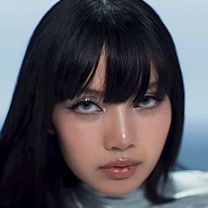 a woman with long black hair wearing a silver shirt and looking at the camera while standing in front of an ocean