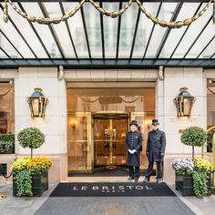 two men standing in front of a hotel entrance with flowers on either side and lights above them