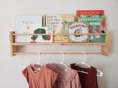 three children's shirts hanging on a wooden shelf with books in the back ground