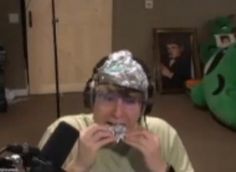 a man wearing a tinfoil hat while eating something in front of him and stuffed animals behind him