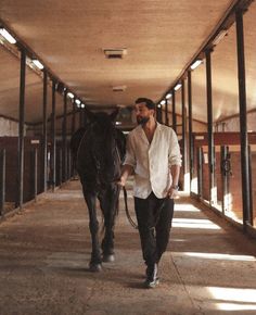 a man walking with a horse down a long hallway lined with stalls on both sides