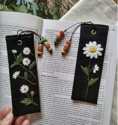 two bookmarks with daisies and leaves on them are hanging from the pages of an open book