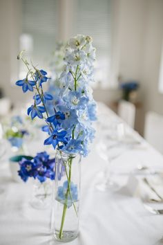 blue and white flowers are in glass vases on a long dining table with silverware