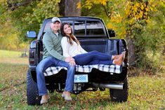 a man and woman sitting on the back of a pick up truck in front of trees