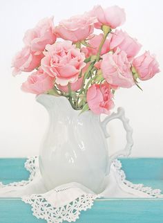 a white vase filled with pink roses on top of a blue table cloth and doily