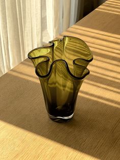 a green glass vase sitting on top of a wooden table next to a white curtain