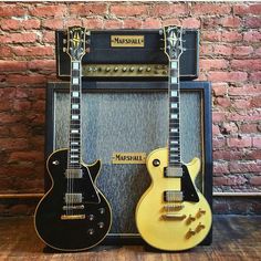 two guitars sitting next to each other in front of a brick wall with an amp