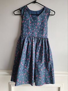 "-This adorable vintage dress is handmade and amazing. The fabric is likely cotton and is navy blue with a simple folk floral pattern in pinks and greens. It is sleeveless with buttons down the back with a sash at the back of waist. There are pockets on the front sides and buttons on the shoulders. Size is: 6x Length: 30.5\" + 1.5\" hem allowance Chest: 16\"" Cotton A-line Sundress For Garden Party, Sleeveless Cotton Sundress With Floral Pattern, Cotton Floral Print Sleeveless Dress For Garden Party, Blue Cotton Sleeveless Sundress, Blue Floral Cotton Sundress, Blue Floral Print Cotton Sundress, Sleeveless Fitted Cotton Floral Dress, Sleeveless Fitted Floral Cotton Dress, Fitted Sleeveless Cotton Floral Dress