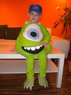 a little boy in a green monster costume sitting on a white table with an orange wall behind him