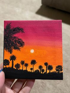 a hand holding up a small painting with palm trees in the background and sunset behind it