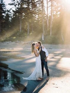 a bride and groom kissing on the beach in front of pine trees at their wedding