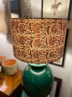 a green vase sitting on top of a wooden table next to a lamp with a paisley print shade