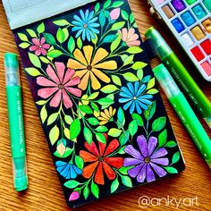 an art journal with flowers painted on it and markers next to the book, pencils and watercolor pens