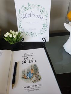 an open book sitting on top of a table next to a pen and flower vase