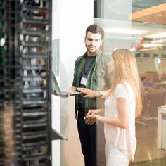 a man and woman standing in front of a server rack talking to each other while holding a laptop computer