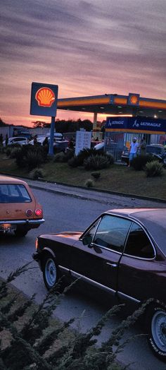 an old car is parked in front of a shell gas station at dusk with the sun setting