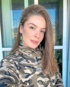 a woman with long brown hair wearing camouflage jacket and earrings standing in front of a door