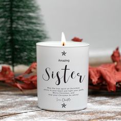 a white candle with the words sister on it sitting next to some red leaves and a small christmas tree