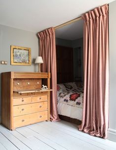 a bed sitting next to a wooden dresser in a bedroom under a window with pink drapes