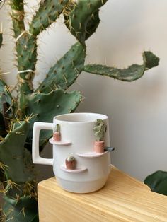 a white coffee cup with pink and green plants on the side sitting on a wooden table next to a cactus