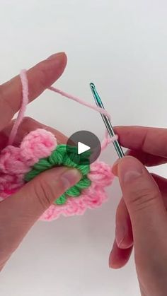 two hands are holding crochet together to make a pink flower with green leaves