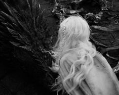 a white doll with long hair sitting on the ground next to dead plants and branches